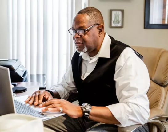 A man sits at a table with his laptop open, tuning into a virtual education session.