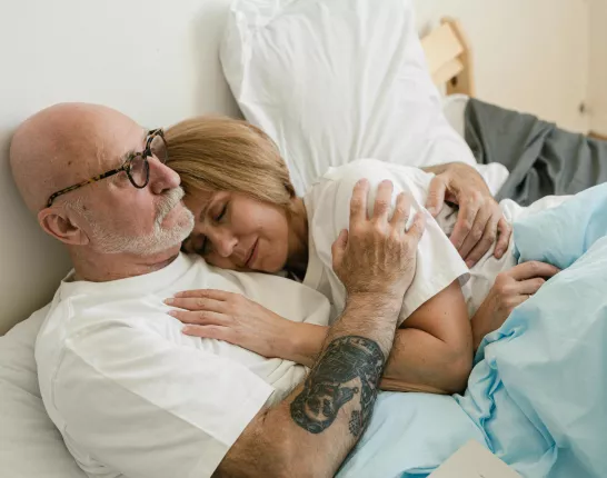 An elderly couple is lying in bed, cuddling closely. The man, wearing glasses and a white shirt, has a tattoo on his arm. The woman rests her head on his chest, eyes closed, with a content expression. They are covered with a light blue blanket.