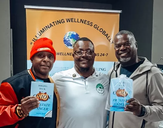 Three men stand smiling in front of a banner that reads "Illuminating Wellness Globally." The man in the center wears a white polo shirt with a green wellness logo, while the two men on either side of him hold up copies of the book “Peeling Back the Onion on Prostate Cancer.” The man on the left wears a red hat and black-and-red varsity jacket, and the man on the right wears a light gray hoodie and sweatpants.
