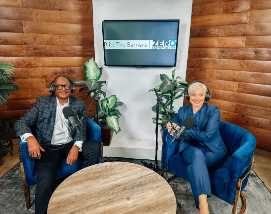 Two individuals are seated in a modern podcast studio. The man on the left wears glasses and a patterned blazer, looking relaxed. The woman on the right is in a navy suit with short platinum blonde hair, smiling warmly. Both wear headphones, ready for recording. A wooden wall and plants add a rustic touch, with a screen displaying "Blitz The Barriers" and "ZERO Prostate Cancer" in the background.