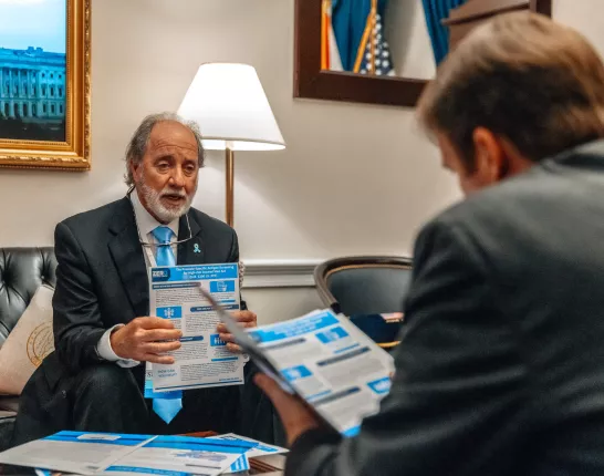 An older man with a white beard, wearing a dark suit and light blue tie, sits in an office setting holding a brochure related to prostate cancer legislation. He appears to be speaking to someone in the foreground whose back is to the camera. The room features official decor including U.S. flags and a framed photo of the Capitol.