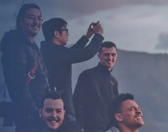 A group of five individuals enjoying an outdoor moment against a mountainous backdrop. The sky is partially cloudy, providing soft lighting. The leftmost person wears a dark jacket with a hood and holds a camera. The second person, slightly behind, is taking a photo with a device. The third person in the middle smiles warmly. The fourth and fifth individuals are also part of the group, sharing the experience.