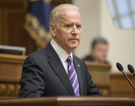 Former President Joe Biden in a dark suit and purple striped tie stands at a podium, delivering a speech in a formal government setting. The background shows a blurred audience and ceremonial flags, indicating a high-level official event.