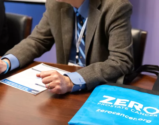 A person in a suit gestures while discussing materials on a table. A blue tote bag with "ZERO PROSTATE CANCER" is prominently displayed, highlighting prostate cancer awareness.