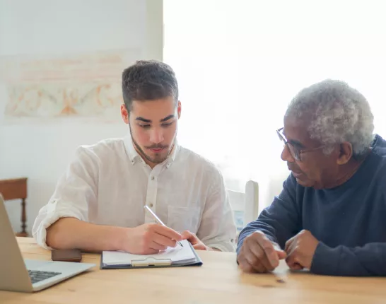 Stock photo of man helping an older man with a notebook and pen sitting at desk