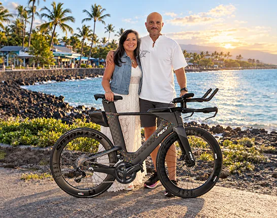 Peter Crescenti and wife standing by bike in front of water and lake and palm trees