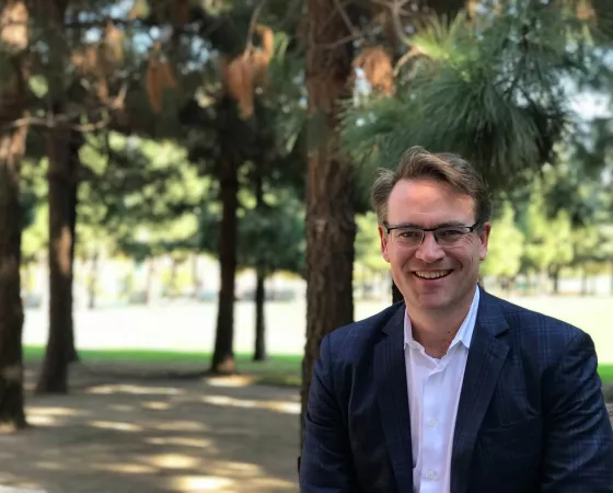 A man wearing smart looking glasses in a suit without a tie sits in shady trees out in the sun