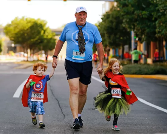 Man holding hands with his two children who are wearing superhero costumes at a Run/Walk