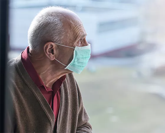 An old white man wearing a mask looking out a window