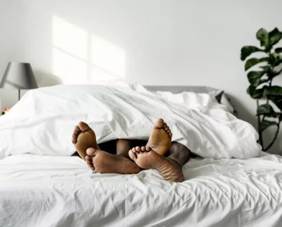 The feet of two Black people sticking out from under the covers of a large bed