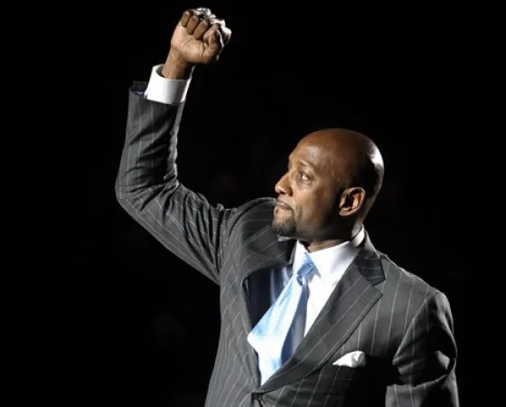 NBA All-Star and Champion, Alonzo Mourning, a black man wearing a grey suit and blue tie with his fist in the air against a black background