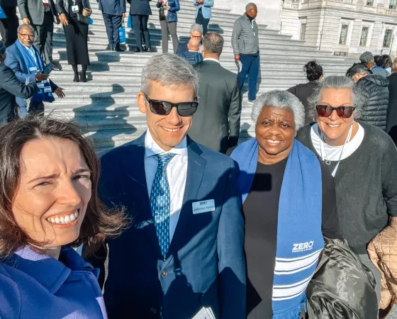 A group of people smiling on the steps of a large building, with some wearing blue scarves that say "ZERO Prostate Cancer." The setting appears to be a formal gathering or event.