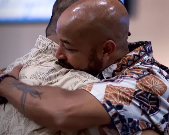 Two men warmly embracing, both wearing colorful, patterned shirts. One has a shaved head and muscular build, the other has short hair and a floral print shirt.
