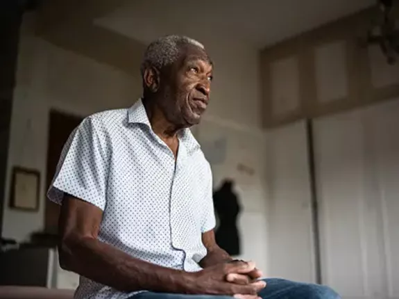 A Black man sitting on the edge of his bed with his hands together