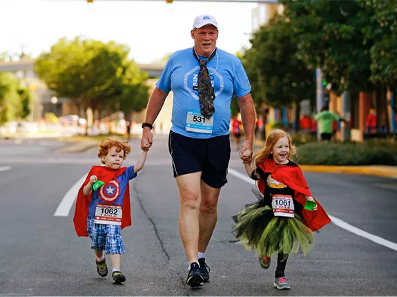 Man holding hands with his two children who are wearing superhero costumes at a Run/Walk