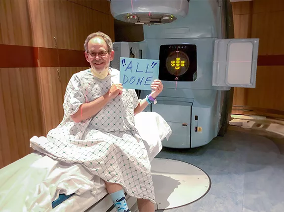 A senior man, Bob Jacobs, sitting in a hospital room with a radiation machine behind him. He's wearing a medical gown and holding a sign that says "All Done"