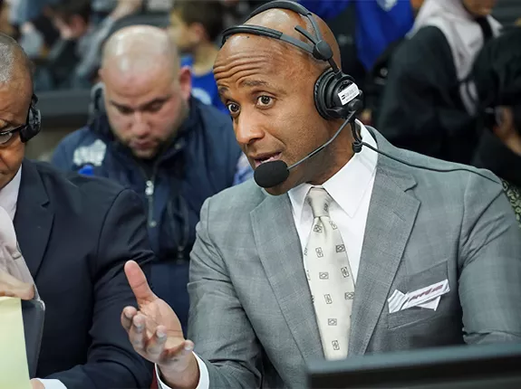 A Black man (Brian Custer) wearing a suite and a headset as an announcer at a sporting event