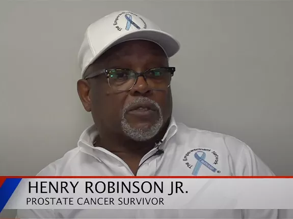 A black man wearing a white polo shirt and white hat, both have a blue ribbon with that says "I Survived". The ribbon is surrounded by text that says "The Empowerment Network". There is also a banner across the bottom that says "Henry Robinson Jr. Prostate Cancer Survivor".
