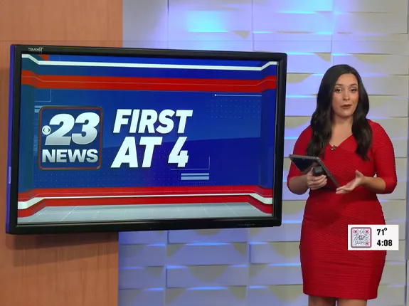 A news anchor wearing a red dress standing next to a television screen that says "23 News First at 4"