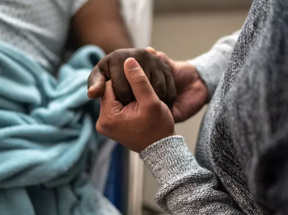 Two black people holding hands; one of them is in a hospital bed
