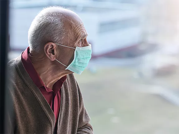 An old white man wearing a mask looking out a window