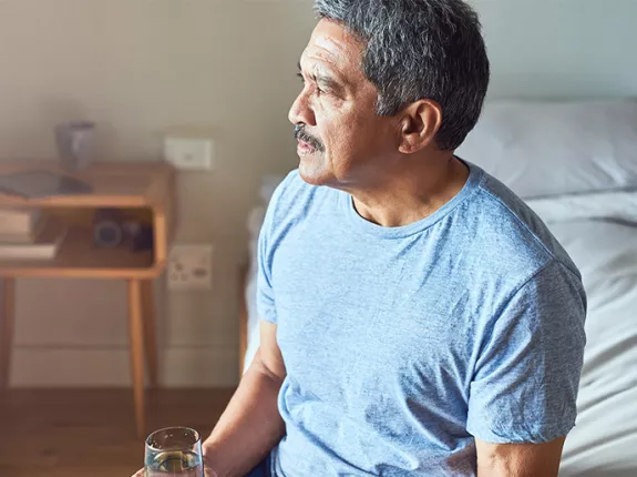 A Hispanic man sitting on the edge of his bed with a glass of water