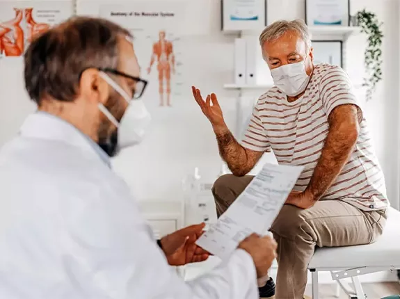 A man sitting on a hospital table wearing a mask while his doctor, also wearing a mask, is reading a paper