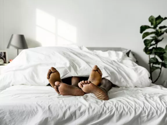 The feet of two Black people sticking out from under the covers of a large bed