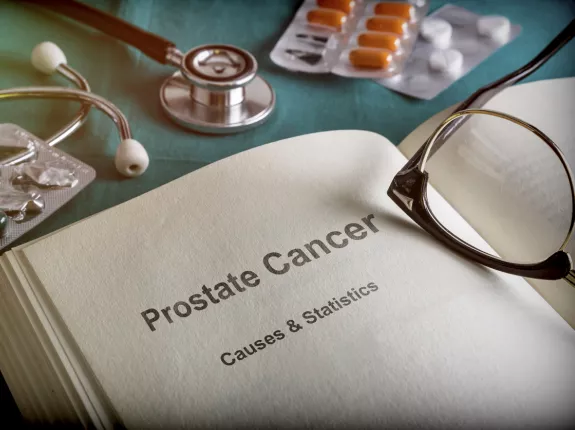 A book opened to a page with text that says "Prostate Cancer Causes & Statistics". There are glasses on the book and prescription pills on the desk.