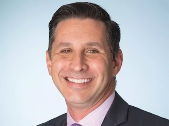 A middle aged man in a suit with dark hair smiling against a light blue background