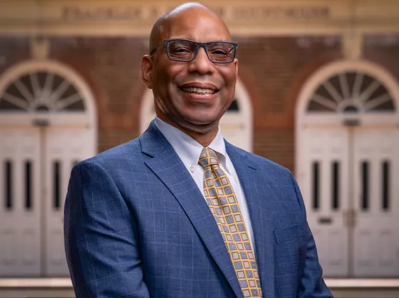 Brian Bragg, Chief Mission Officer, a Black man in a blue suit and yellow checkered tie standing in front of a brick building