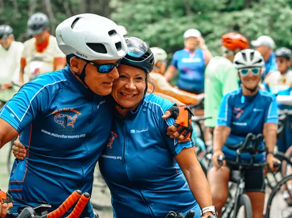 An older couple in cycling jerseys and helmets embracing while surround by many onlooking cyclists