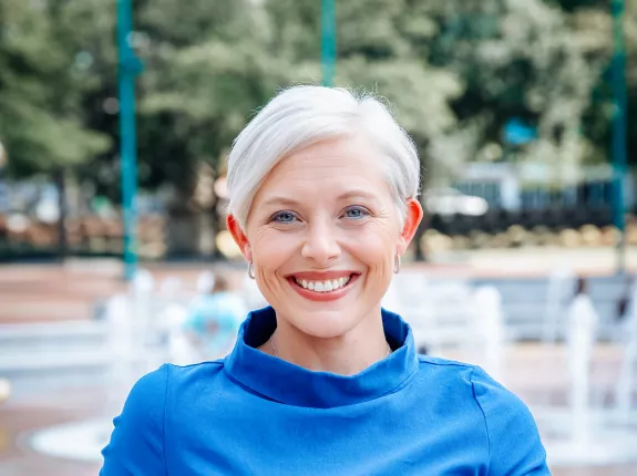 Blonde woman with short hair wearing a blue dress standing in front of a fountain