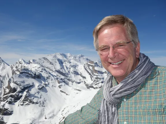 A blonde man with glasses standing in front of the Swiss Alps