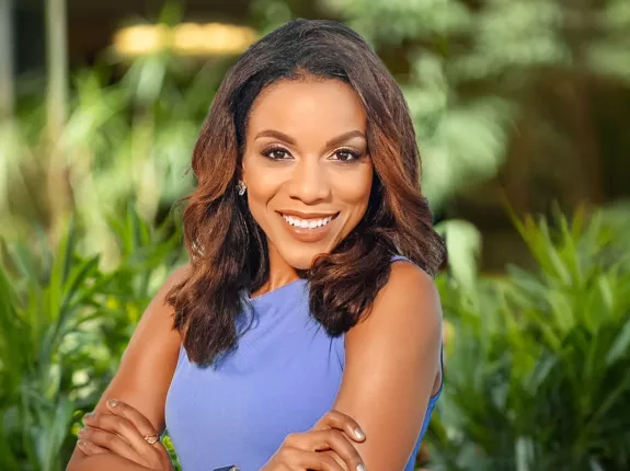 A Black woman in a purple top standing with her arms crossed and smiling in front of green foliage