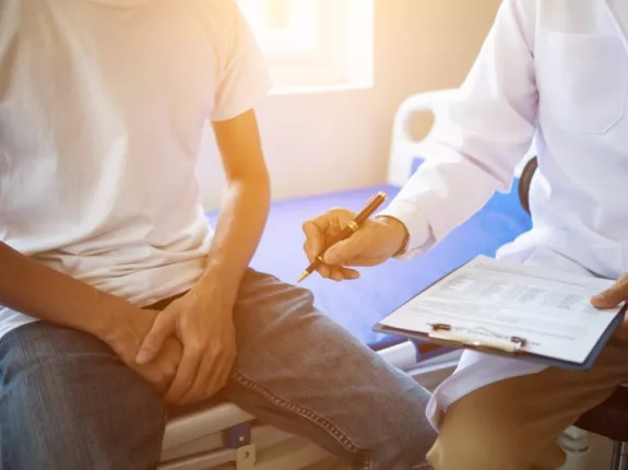 A shot of a man wearing a white tshirt and jeans sitting on a doctors table with his hands in his lap and a doctor sitting by the table on a stool holding a clipboard