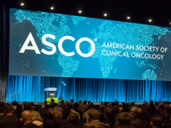 A large conference hall with a wide stage featuring a prominent display screen showing the ASCO (American Society of Clinical Oncology) logo and text in white against a turquoise world map background. The stage is framed by blue-lit curtains and lighting. The audience area is filled with attendees seated in rows, viewed from behind. The overall lighting creates a professional, dramatic atmosphere typical of major medical conferences.