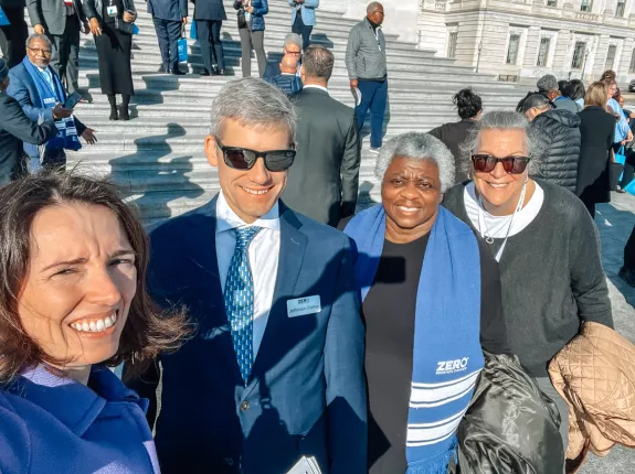 A group of people smiling on the steps of a large building, with some wearing blue scarves that say "ZERO Prostate Cancer." The setting appears to be a formal gathering or event.