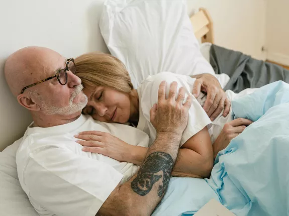 An elderly couple is lying in bed, cuddling closely. The man, wearing glasses and a white shirt, has a tattoo on his arm. The woman rests her head on his chest, eyes closed, with a content expression. They are covered with a light blue blanket.