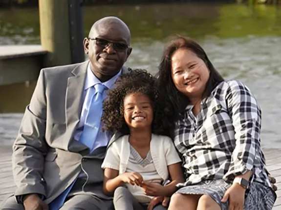 A man in a gray suit and sunglasses, a smiling young girl in a light outfit, and a woman in a checkered shirt sit together on a wooden deck by the water, exuding warmth and happiness.