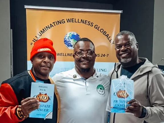 Three men stand smiling in front of a banner that reads "Illuminating Wellness Globally." The man in the center wears a white polo shirt with a green wellness logo, while the two men on either side of him hold up copies of the book “Peeling Back the Onion on Prostate Cancer.” The man on the left wears a red hat and black-and-red varsity jacket, and the man on the right wears a light gray hoodie and sweatpants.