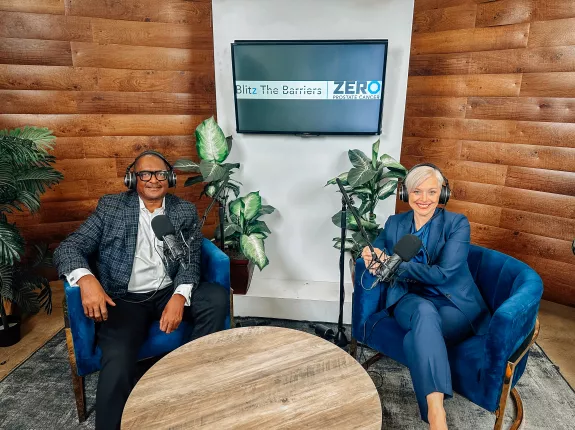 Two individuals are seated in a modern podcast studio. The man on the left wears glasses and a patterned blazer, looking relaxed. The woman on the right is in a navy suit with short platinum blonde hair, smiling warmly. Both wear headphones, ready for recording. A wooden wall and plants add a rustic touch, with a screen displaying "Blitz The Barriers" and "ZERO Prostate Cancer" in the background.