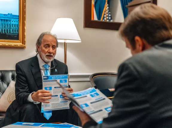 An older man with a white beard, wearing a dark suit and light blue tie, sits in an office setting holding a brochure related to prostate cancer legislation. He appears to be speaking to someone in the foreground whose back is to the camera. The room features official decor including U.S. flags and a framed photo of the Capitol.