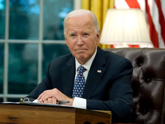 Former President Joe Biden in a suit sits at a desk in a formal office, looking serious. An American flag and lamp are in the background.