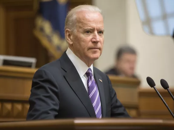 Former President Joe Biden in a dark suit and purple striped tie stands at a podium, delivering a speech in a formal government setting. The background shows a blurred audience and ceremonial flags, indicating a high-level official event.