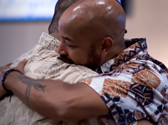 Two men warmly embracing, both wearing colorful, patterned shirts. One has a shaved head and muscular build, the other has short hair and a floral print shirt.