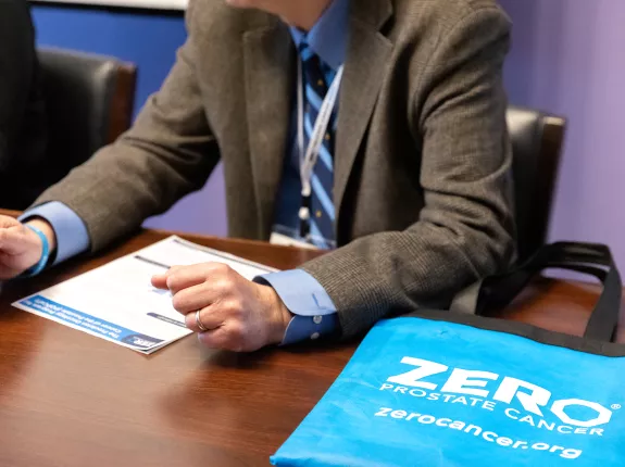A person in a suit gestures while discussing materials on a table. A blue tote bag with "ZERO PROSTATE CANCER" is prominently displayed, highlighting prostate cancer awareness.