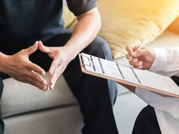 Image of hands of a patient and doctor. The doctor is holding a clipboard and appears to be consulting with the patient.