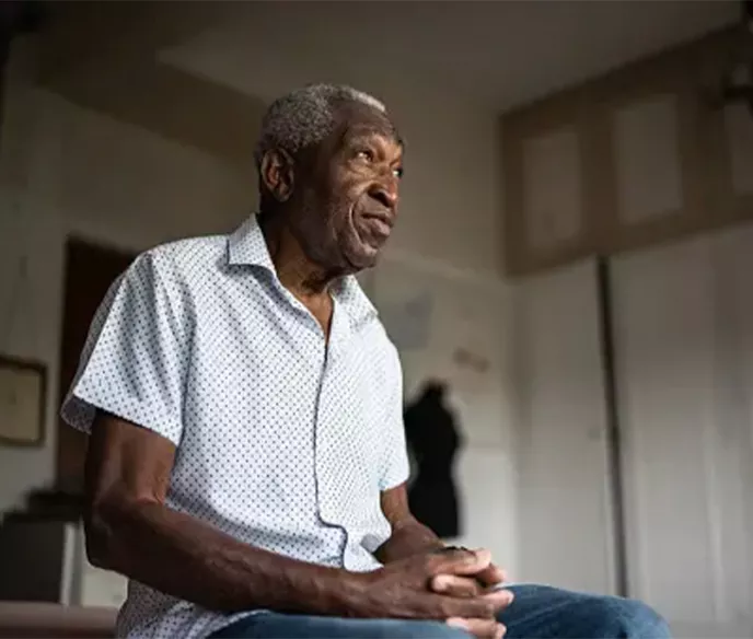 A Black man sitting on the edge of his bed with his hands together