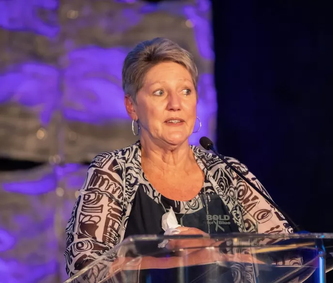 A woman with short blond hair and teary eyes speaking at an event